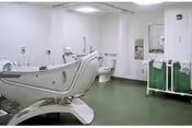 A spacious bathroom in a nursing and rehab facility featuring a large white bathtub with a reclining chair design, a toilet, a sink with a mirror above it, a green floor, and a green cart with towels. The walls are white and there is a shower curtain near the bathtub.