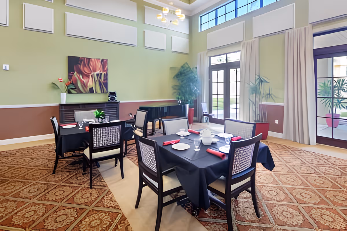 Dining room with tables set with black tablecloths, red napkins, and chairs beneath tall windows and high ceilings.