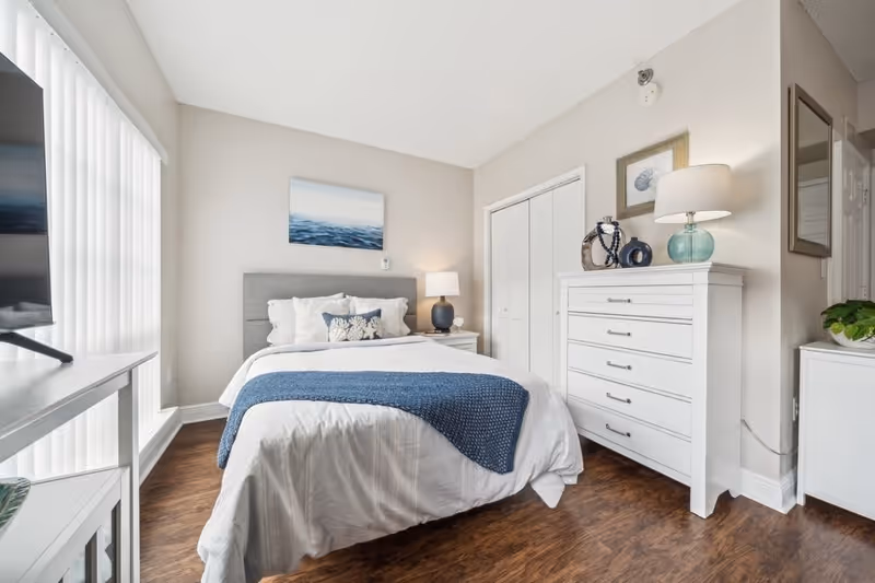 A bright and clean bedroom with a neatly made bed featuring white bedding and a blue throw blanket. The room has wooden flooring, a white dresser with decorative items and a lamp on top, a closet with white doors, and a large window with vertical blinds allowing natural light to enter. A flat-screen TV is positioned on a white stand opposite the bed.