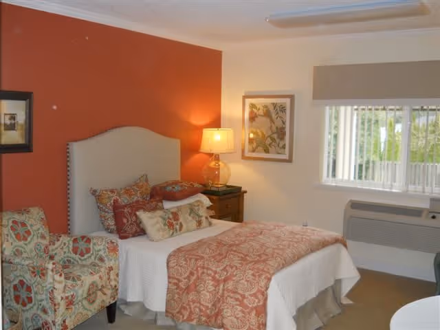 A cozy bedroom with a bed featuring a beige upholstered headboard, white bedding, and a red patterned throw blanket. There are multiple decorative pillows on the bed. To the left of the bed is a floral patterned armchair. A wooden nightstand with a lamp and a framed picture on the wall are visible. A window with vertical blinds lets in natural light, and an air conditioning unit is installed below the window.