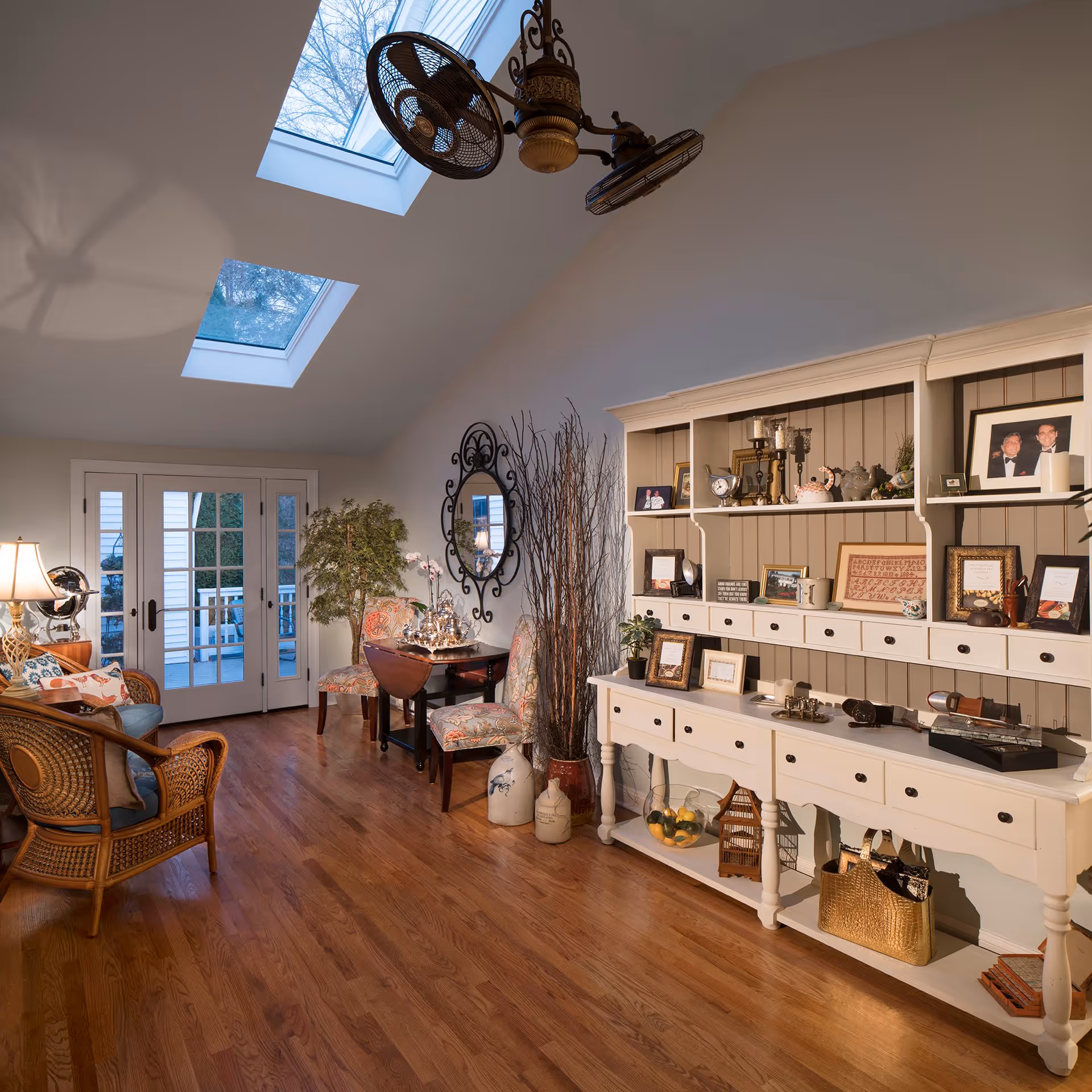 A cozy living room area with wooden flooring, a white cabinet filled with framed photos and decorative items, two floral upholstered chairs next to a small wooden table, a wicker chair with cushions, a large round decorative mirror on the wall, and two skylights in the ceiling letting in natural light. French doors lead to an outdoor deck.