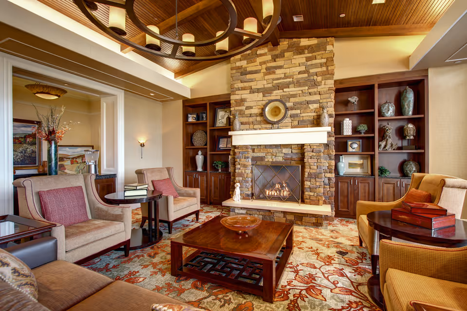 A cozy living room with a stone fireplace in the center, flanked by built-in wooden shelves filled with decorative items. The room features comfortable armchairs with red cushions, a wooden coffee table, and a patterned carpet. The ceiling has wooden paneling and a circular chandelier with warm lighting.