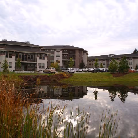 Exterior view of a senior living facility with multiple buildings surrounding a small pond. The buildings are three to four stories tall with balconies and windows. There is a parking lot with several cars and landscaped greenery including trees and grass around the pond.