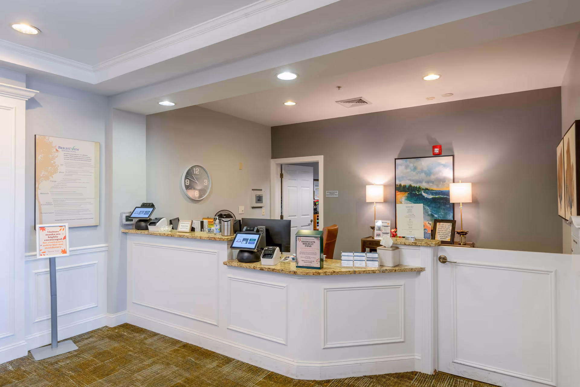 Reception area of Brightview South River Senior Assisted Living & Memory Care with a curved white front desk, two computer monitors, informational signs, a wall clock, and a painting on the wall behind the desk illuminated by two table lamps.