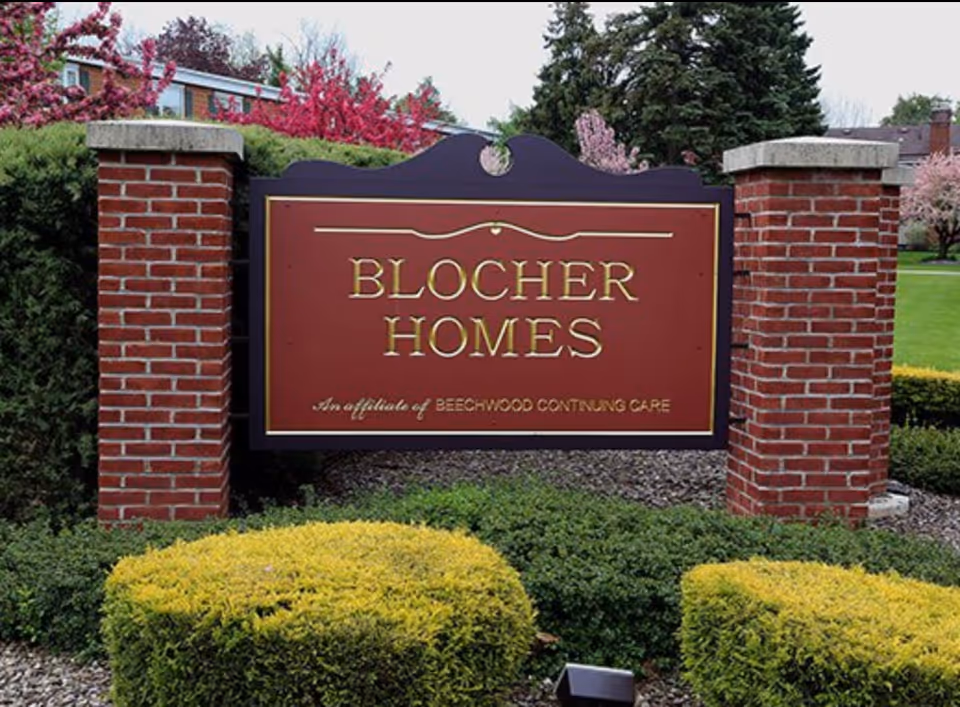 A large maroon and gold sign reading 'Blocher Homes, An affiliate of Beechwood Continuing Care' mounted between two brick pillars with greenery and flowering trees in the background.