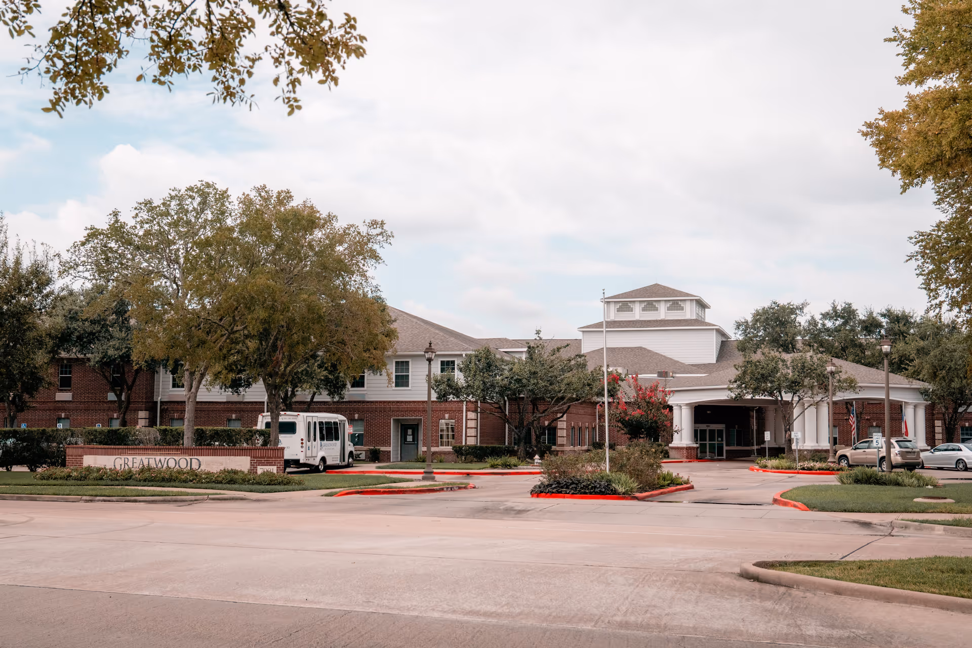 Front exterior of the Greatwood at Sugar Land senior living building with driveway, landscaping, and parked vehicles.