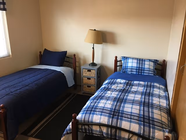 Two single beds with blue bedding separated by a small nightstand and lamp in a simple bedroom.