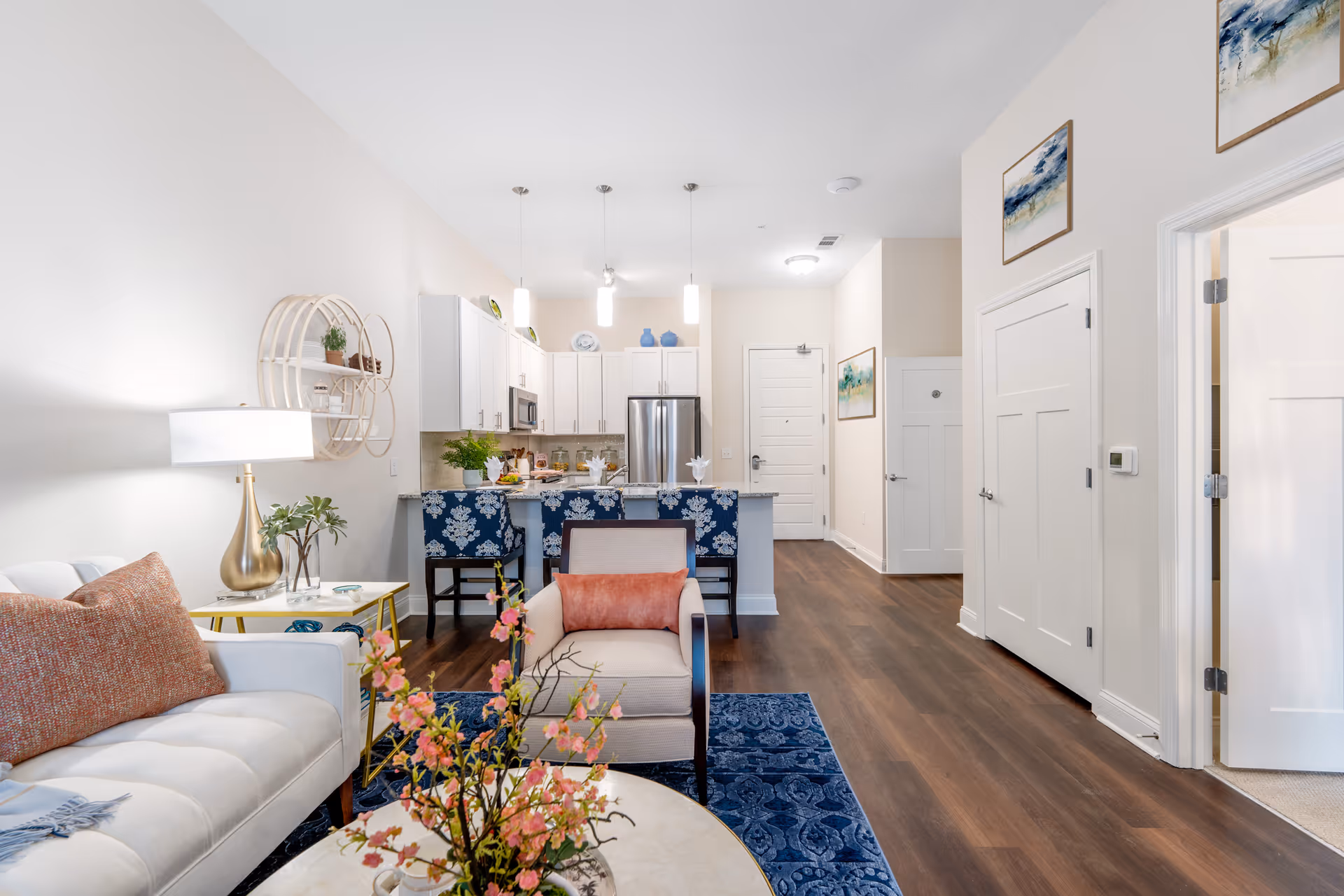 Bright and modern open-concept living area featuring a white sofa with a rust-colored pillow, a beige armchair with a matching pillow, a round coffee table with a floral arrangement, and a blue patterned rug. The space includes a kitchen with white cabinets, stainless steel appliances, and a breakfast bar with three blue floral-patterned stools. The room has wood flooring, white walls, and decorative wall art.