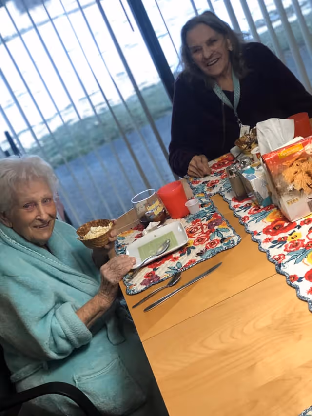 Two elderly women sitting at a dining table with colorful floral placemats. One woman is wearing a light blue robe and holding a waffle bowl with food, while the other woman is smiling and wearing a dark sweater. The table has various items including a red cup, a glass, a box of crackers, and utensils. Vertical blinds and a window are visible in the background.