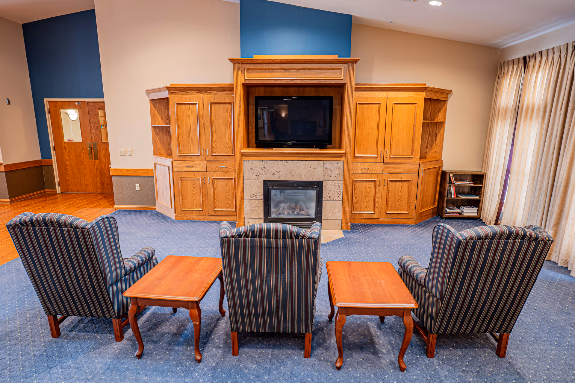 A cozy common area with three striped armchairs arranged in front of a wooden entertainment center featuring a flat-screen TV and a fireplace. The room has blue carpet, wooden side tables between the chairs, beige walls, and large windows with curtains on the right side.