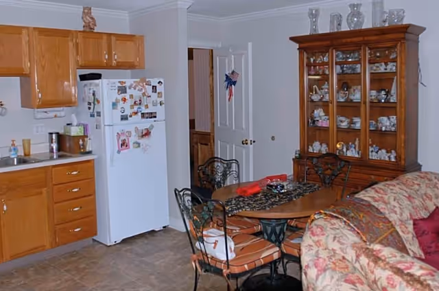 Interior view of a senior living facility showing a kitchen area with wooden cabinets, a white refrigerator with magnets, a sink, and a dining area with a round wooden table and four chairs. There is a wooden china cabinet filled with decorative items and a floral-patterned sofa partially visible in the foreground.