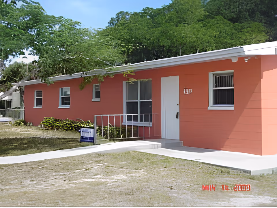 Single-story coral-colored building front with a white door, windows, a small porch railing, and a grassy yard.