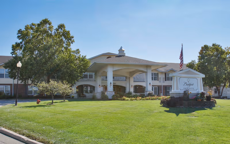 Front exterior view of Bickford of Bloomington senior living facility with a large covered entrance, well-maintained lawn, trees, and a sign displaying the facility name alongside an American flag.