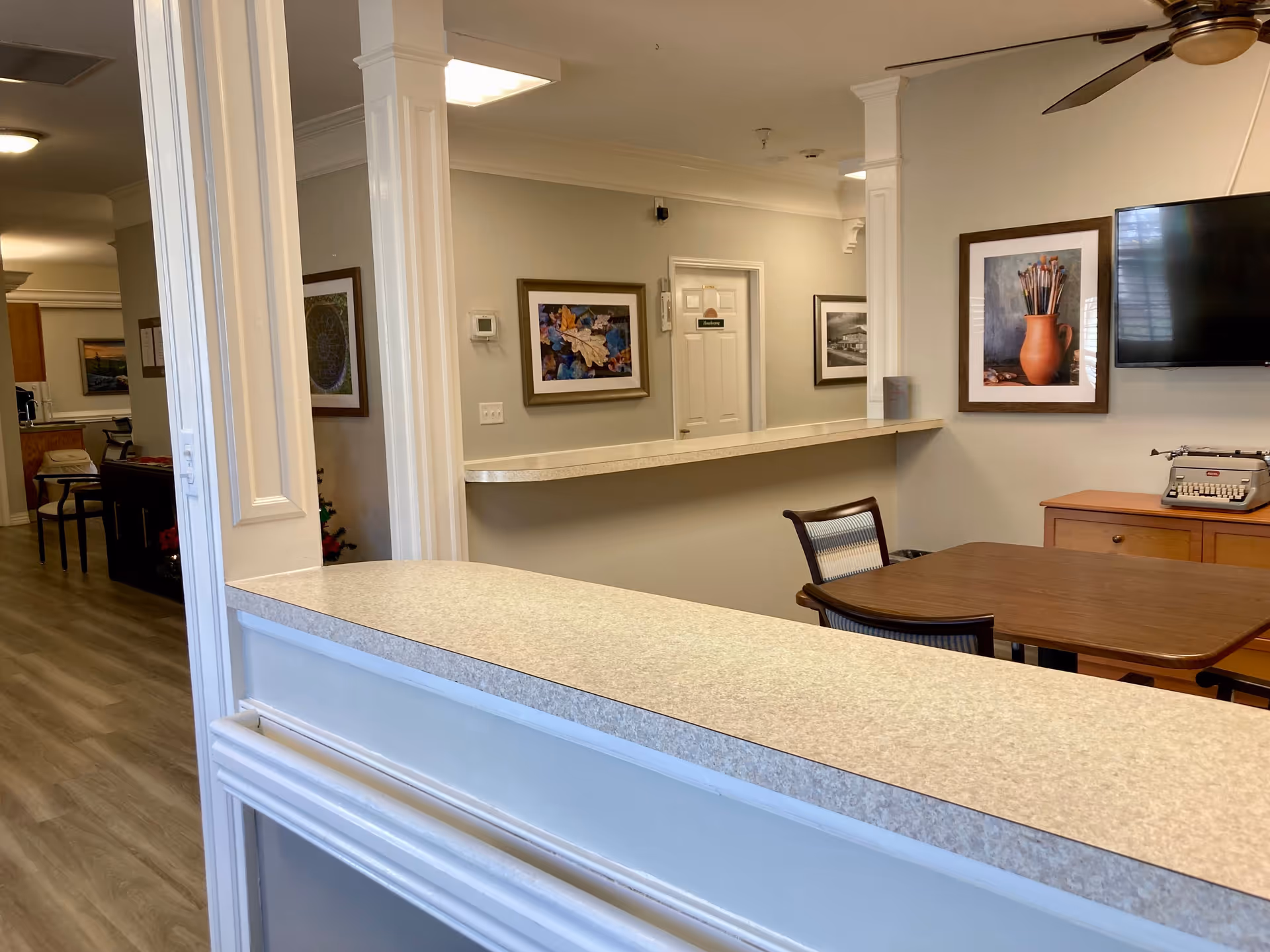 Interior view of a senior living facility showing a reception or common area with a countertop in the foreground, a wooden table with chairs, framed artwork on the walls, a wall-mounted TV, and a typewriter on a wooden cabinet. The hallway leads to other rooms with closed doors and additional artwork.