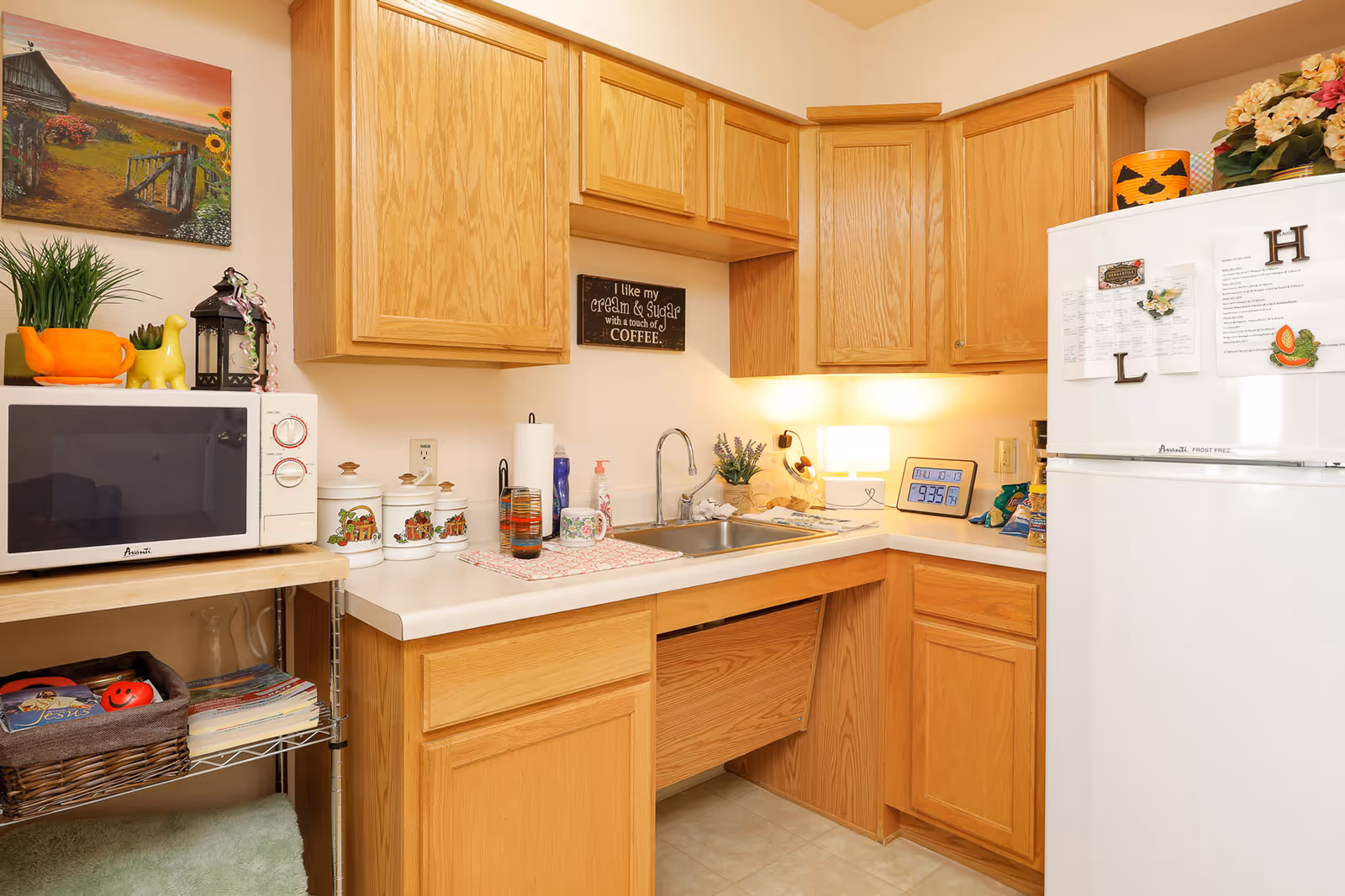 A small kitchen area with wooden cabinets, a white countertop, a stainless steel sink, a white microwave on a metal shelf, and a white refrigerator decorated with magnets and papers. The countertop has various items including a glass, a mug, a paper towel holder, a small lamp, and a digital clock. There is a painting on the wall and some decorative plants and items on the microwave and refrigerator.