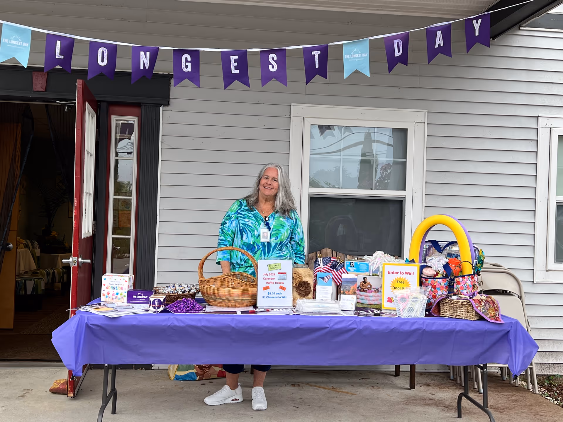 A woman with gray hair wearing a blue and green patterned shirt stands behind a table covered with a purple tablecloth. The table displays various items including baskets, raffle tickets, and promotional materials. Above her, a banner reads 'LONGEST DAY'. The setting is outside a building with gray siding and a red door.