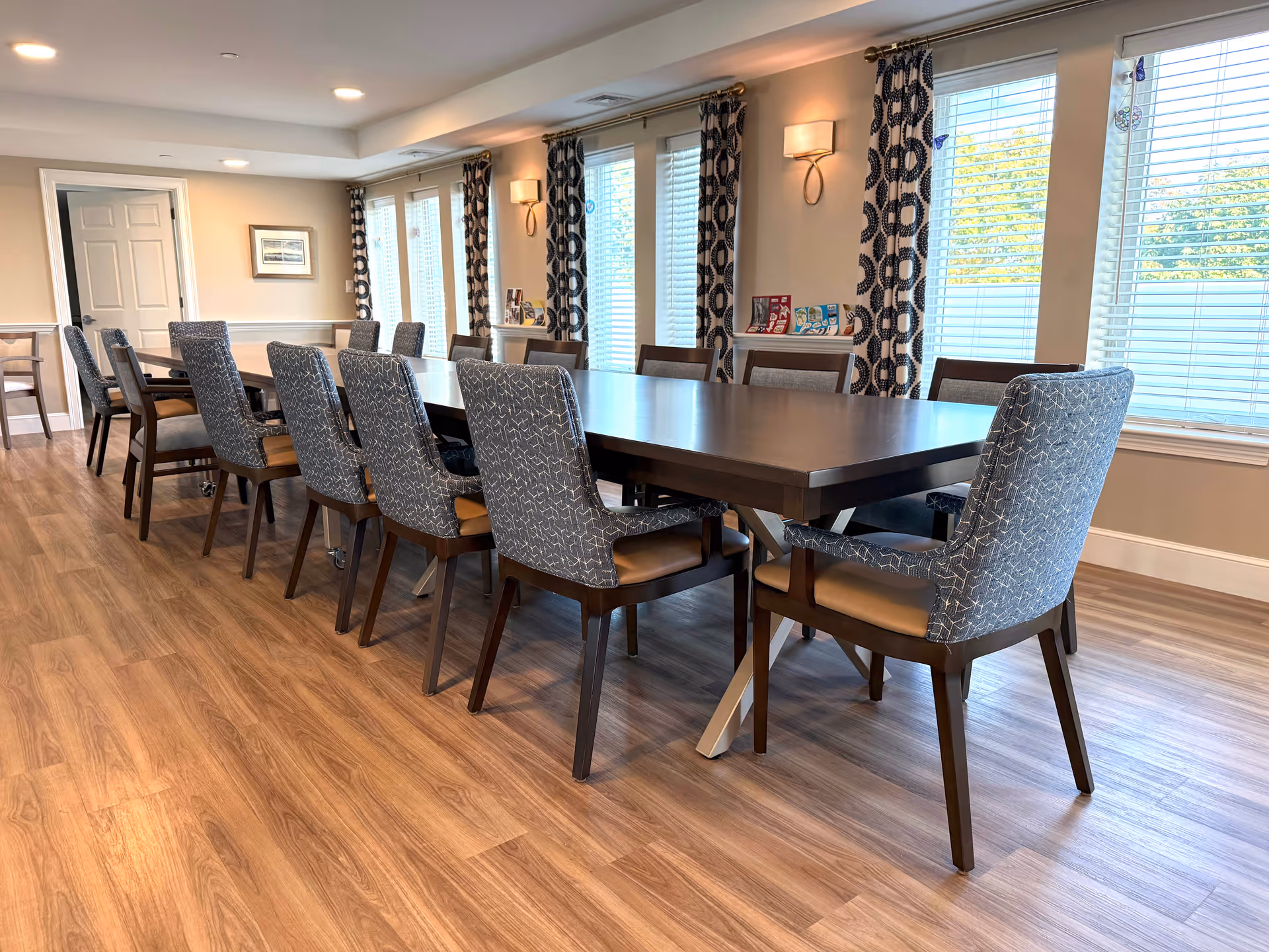 Long dining table surrounded by upholstered chairs in a bright dining room with wood flooring and windows with patterned curtains.