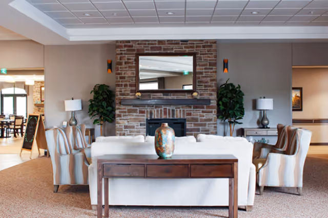 A cozy living room area in an assisted living facility featuring a white sofa and two striped armchairs arranged around a brick fireplace with a large mirror above it. There are two potted plants on either side of the fireplace and two table lamps on side tables. A wooden console table with a decorative vase is placed behind the sofa.