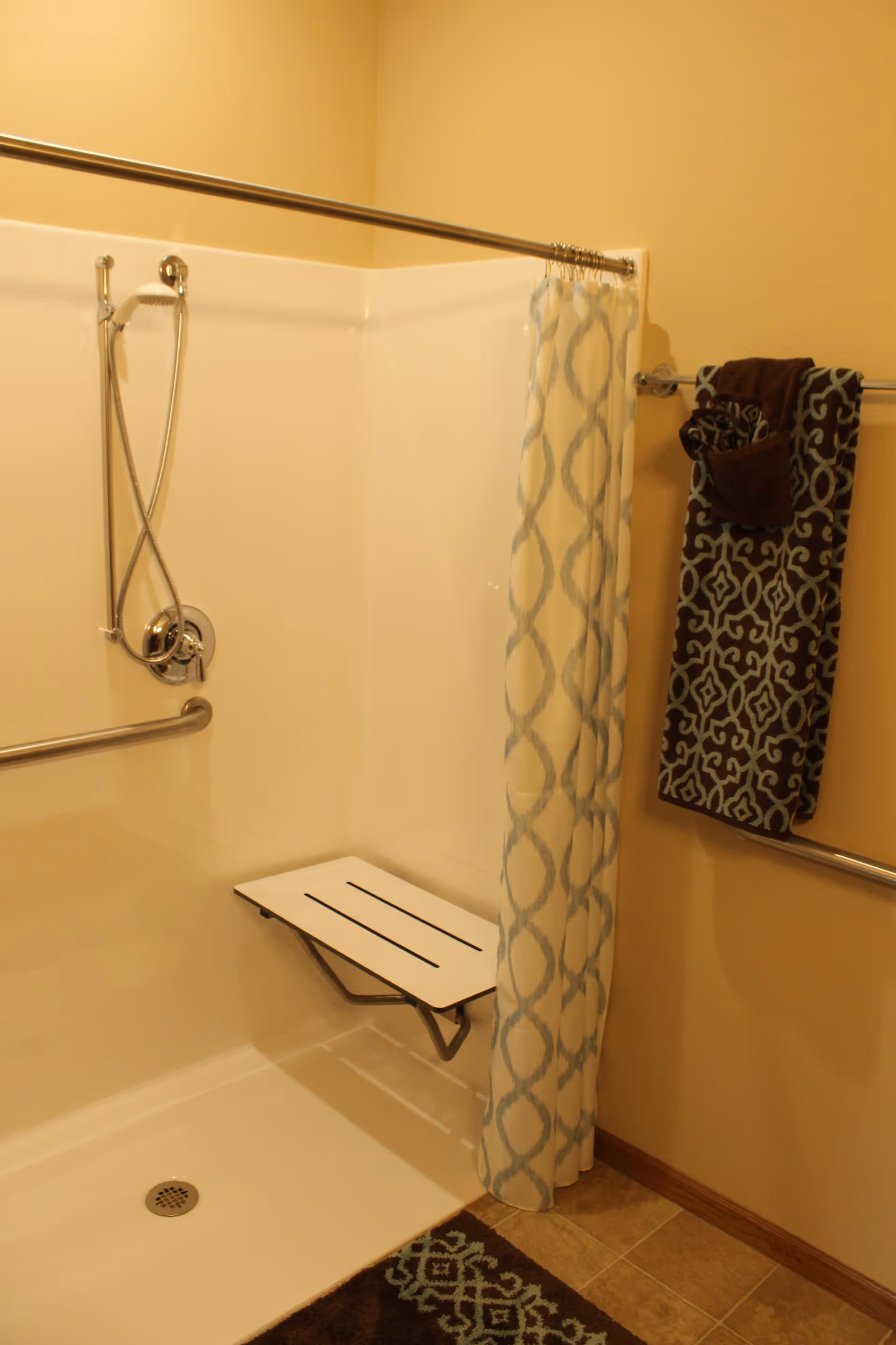 A bathroom shower area with a white fold-down seat attached to the wall, a handheld showerhead, and a silver grab bar. There is a patterned shower curtain partially drawn to the side, and a towel rack on the wall holding a patterned towel and a smaller brown towel. The floor has a brown and blue patterned bath mat.