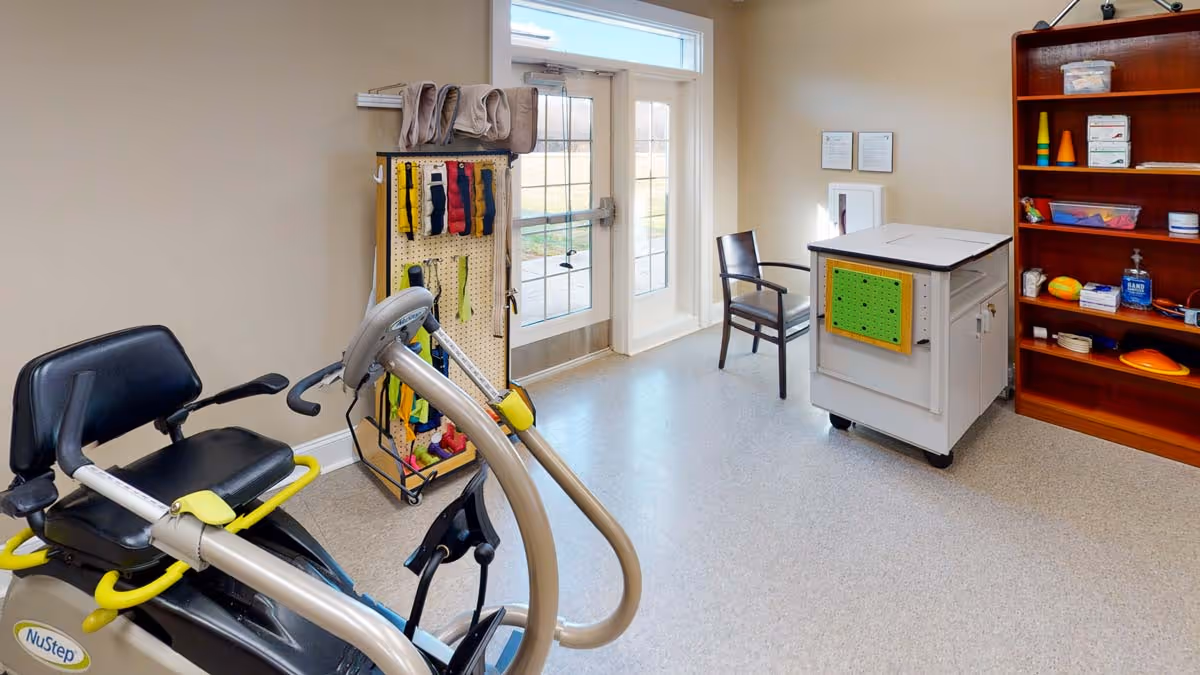 A bright therapy or exercise room with a NuStep exercise machine in the foreground, a pegboard with various exercise bands and equipment hanging on it, a chair, a small rolling cart with a pegboard attached, and a wooden shelf with therapy supplies and hand sanitizer. A glass door leads outside, allowing natural light into the room.