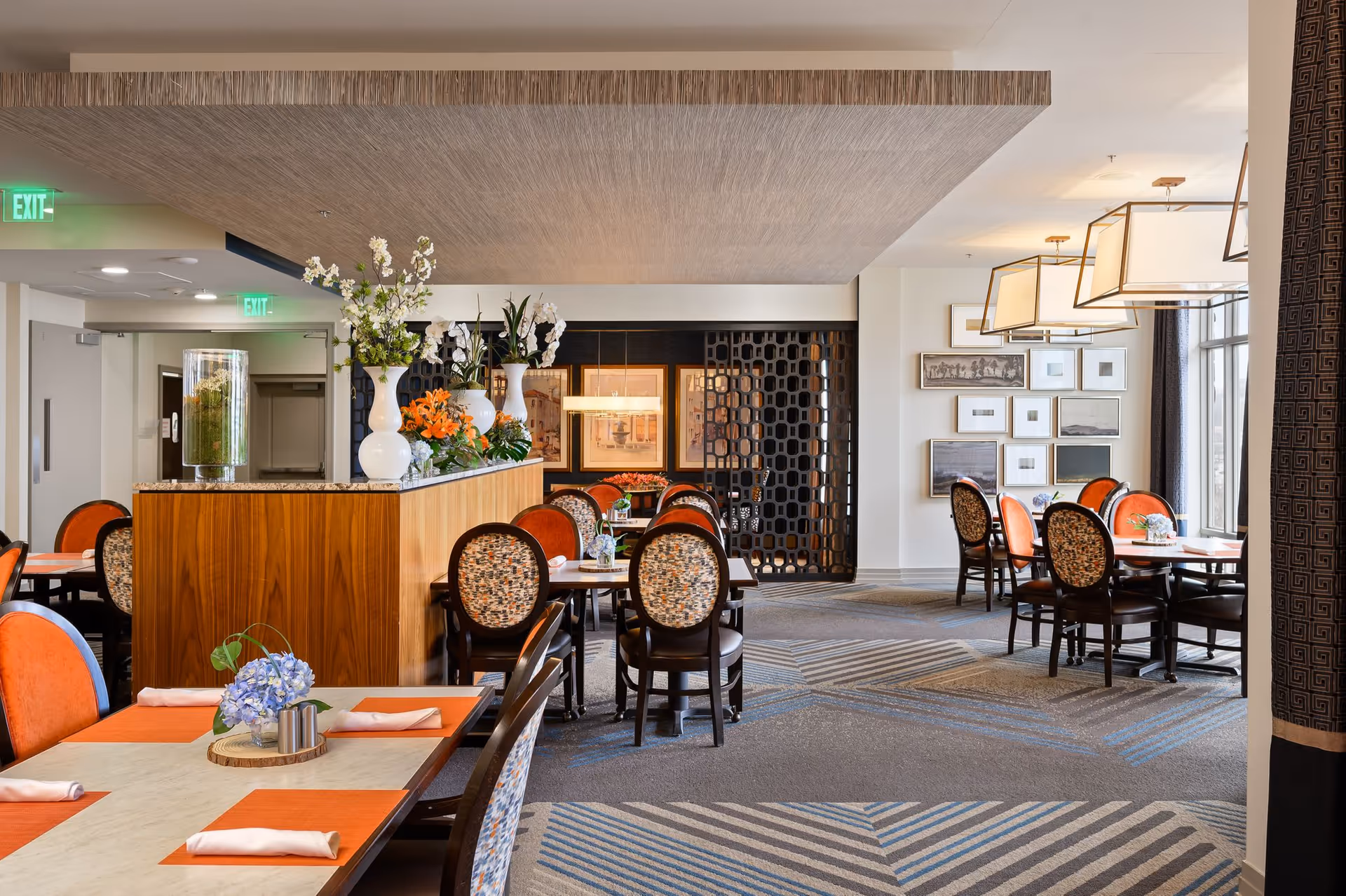 A bright and elegant dining room in a senior living facility with tables set with orange placemats, white napkins, and small floral centerpieces. The room features upholstered chairs with patterned backs and orange seats, decorative vases with flowers on a wooden divider, framed artwork on the walls, and large windows letting in natural light.
