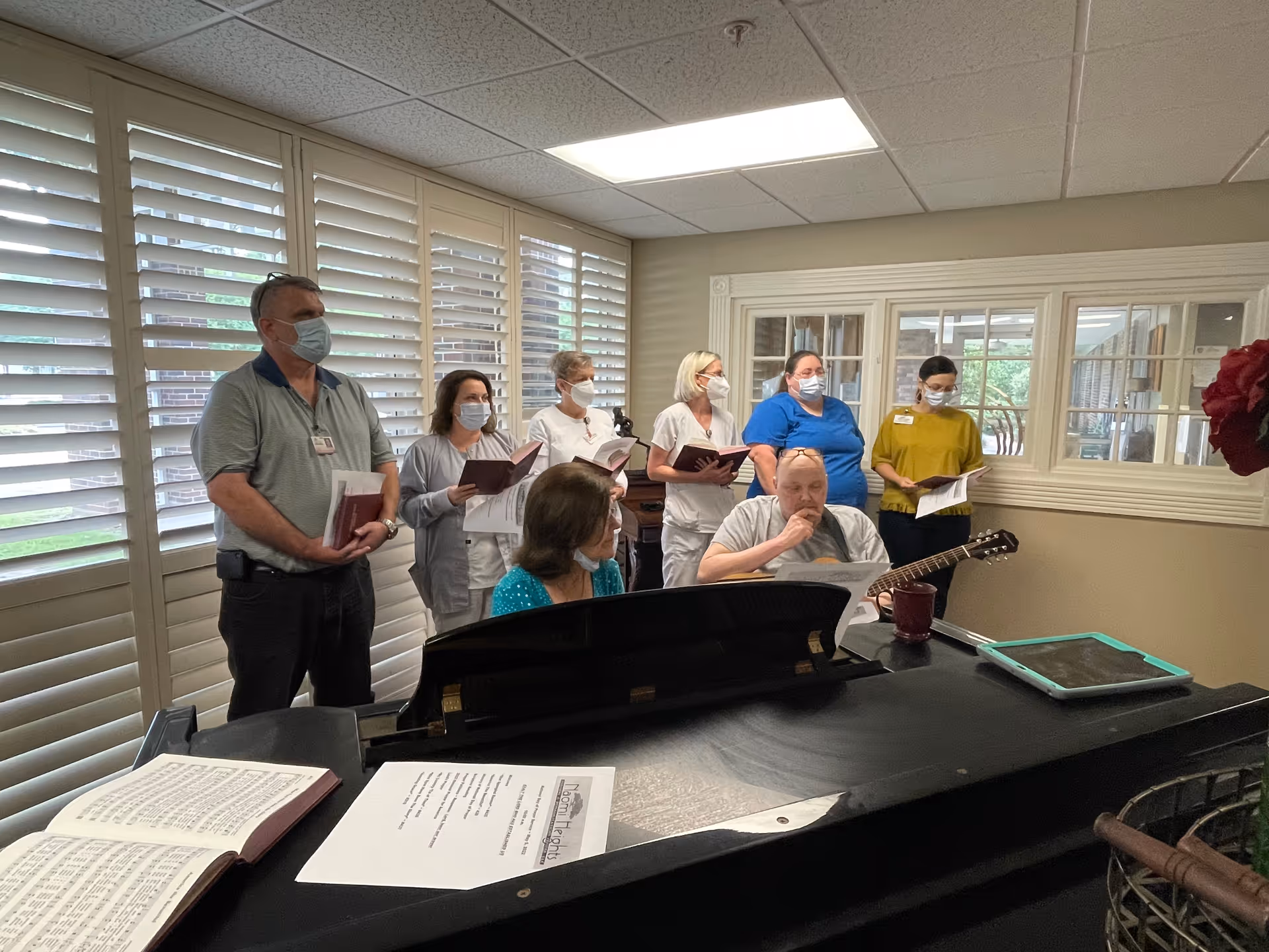 A group of seven adults wearing face masks standing and sitting around a piano in a room with large windows covered by white shutters. Some individuals are holding songbooks, and one person is playing a guitar. The setting appears to be a communal or activity room in a nursing or rehabilitation center.
