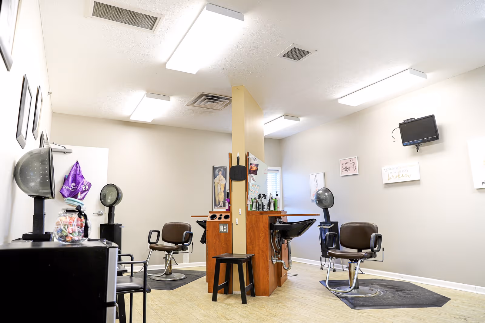 Interior view of a hair salon area with two salon chairs, hair dryers, a wash basin, and various hair care products. The room has light-colored walls with framed pictures and a small wall-mounted TV. The floor is light wood, and the ceiling has fluorescent lighting.