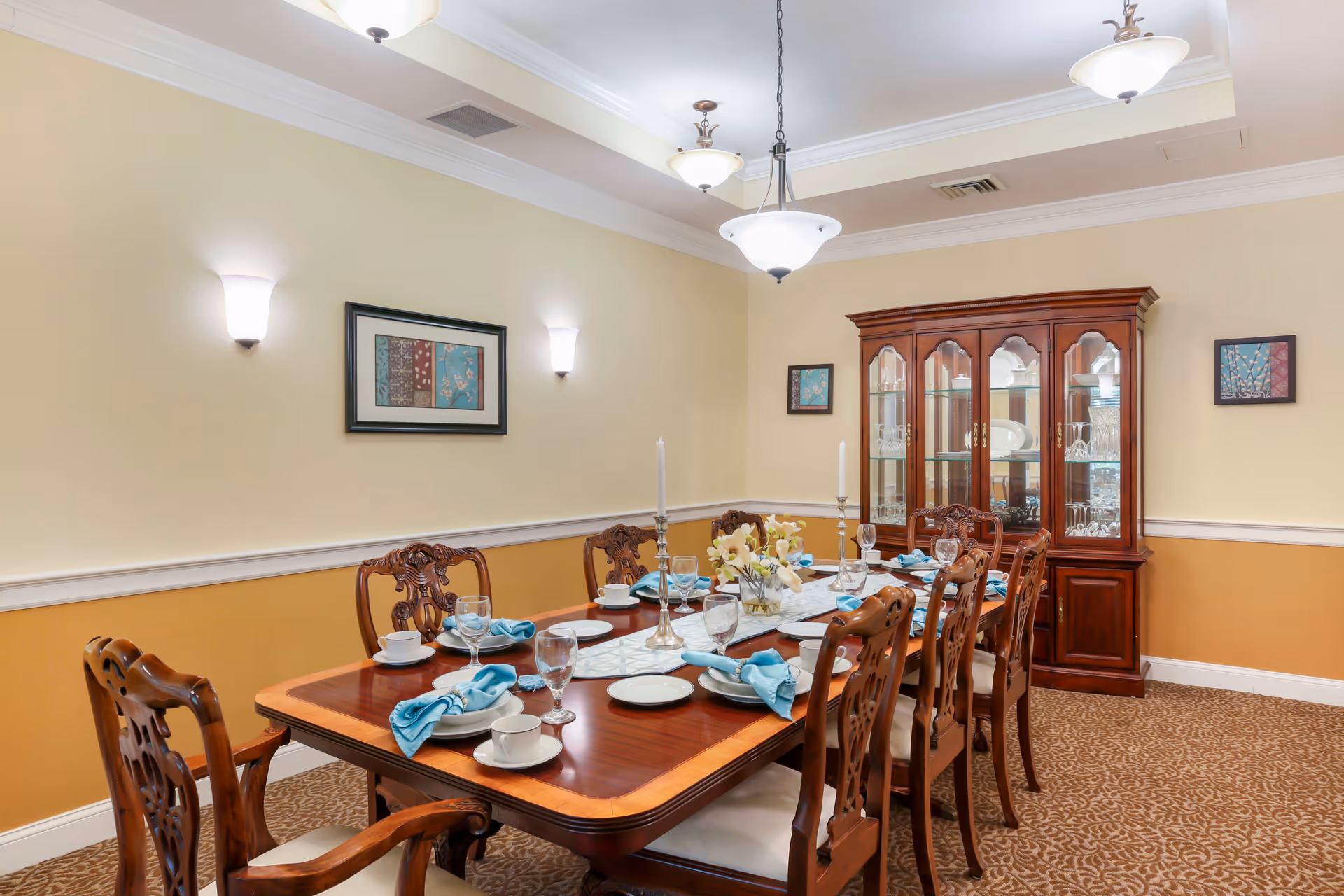 A formal dining room with a long wooden table set for eight people. The table is arranged with white plates, cups, glasses, and blue cloth napkins. There are two tall candlesticks and a floral centerpiece on the table. The room has beige and light yellow walls with three framed artworks and a wooden china cabinet filled with glassware. The ceiling features recessed lighting and hanging light fixtures.