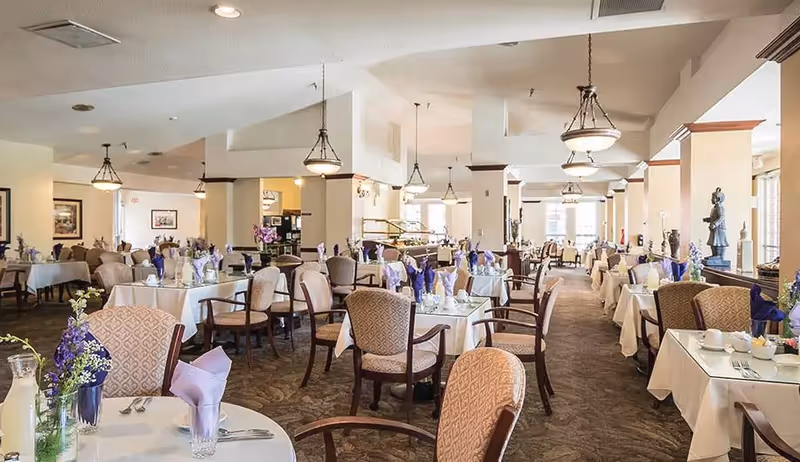 Bright dining room with multiple set tables, upholstered chairs, floral centerpieces, and hanging light fixtures.