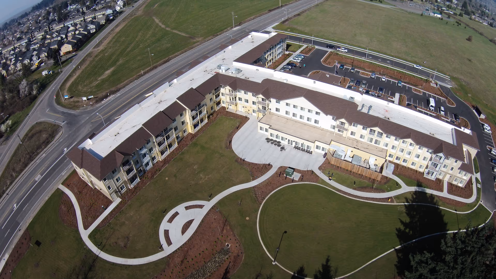 Aerial view of Volante Senior Living of Eugene, showing a large U-shaped building with a brown roof and light-colored walls. The building is surrounded by well-maintained lawns, curved walking paths, and a parking lot with several cars. The surrounding area includes open green fields and a nearby residential neighborhood.