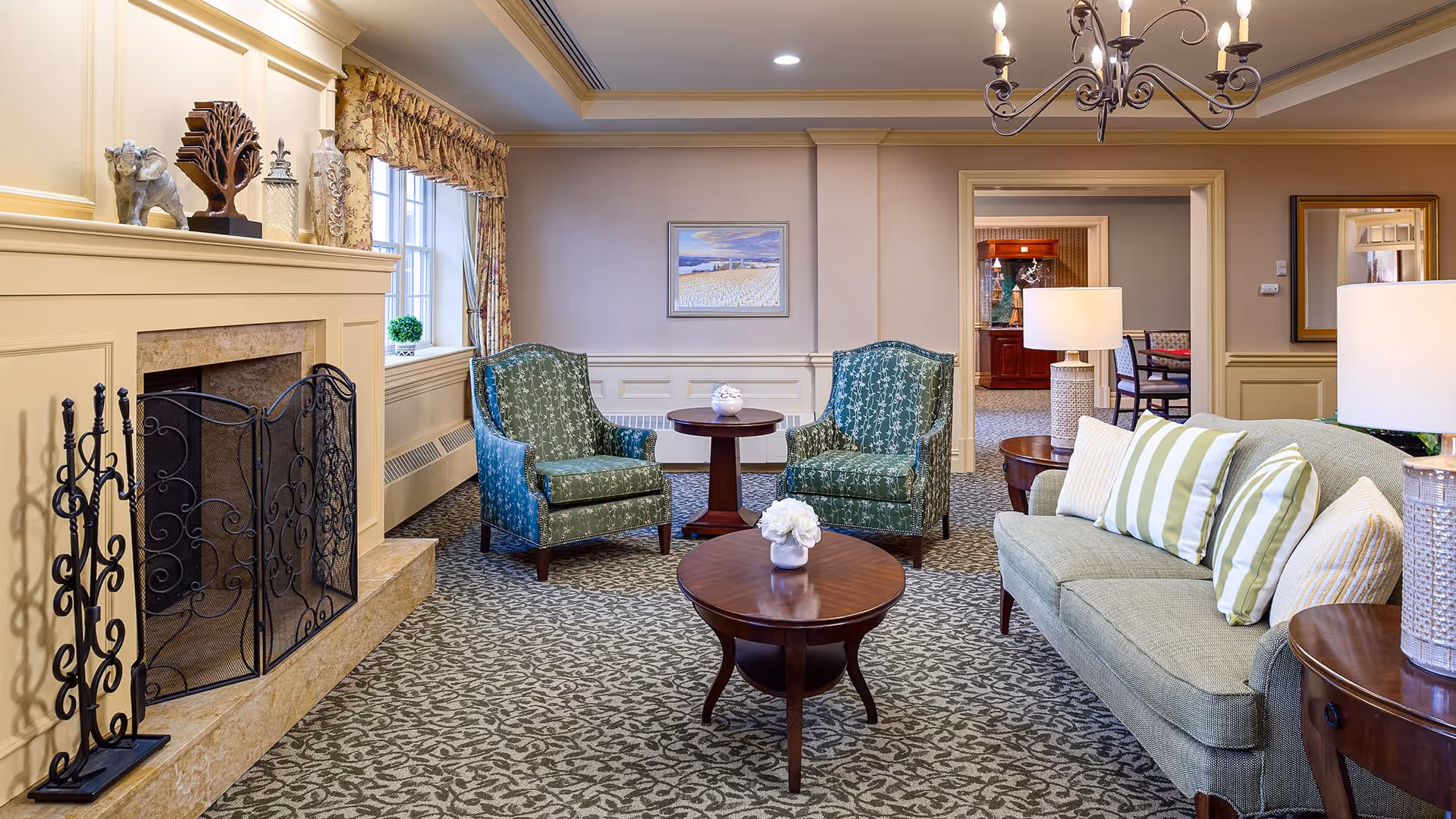 A cozy living room area in a senior living facility featuring a beige fireplace with decorative items on the mantel, two green patterned armchairs, a green sofa with striped and patterned pillows, two round wooden coffee tables with white floral arrangements, and two table lamps. The room has patterned carpet, beige walls with wainscoting, a window with floral curtains, and a chandelier hanging from the ceiling.
