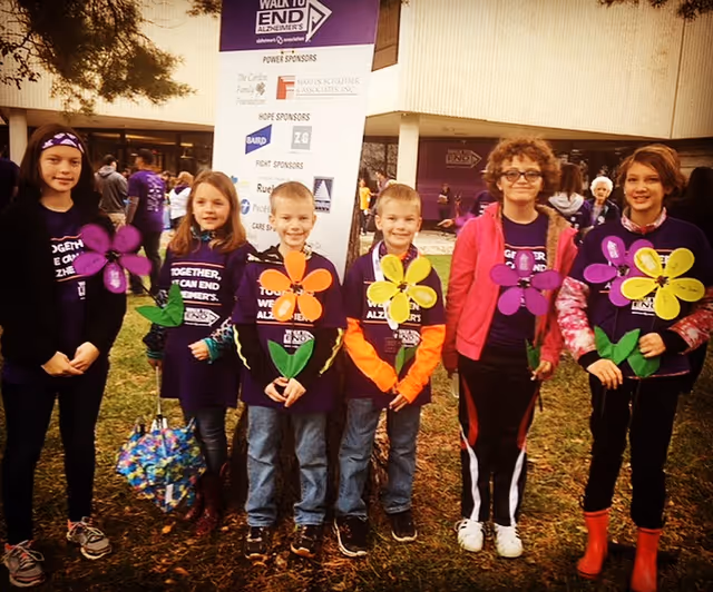 Six children standing outdoors on grass in front of a building, each holding colorful flower-shaped signs. They are wearing purple shirts with text about ending Alzheimer's, and there is a large sign behind them listing sponsors for a Walk to End Alzheimer's event.