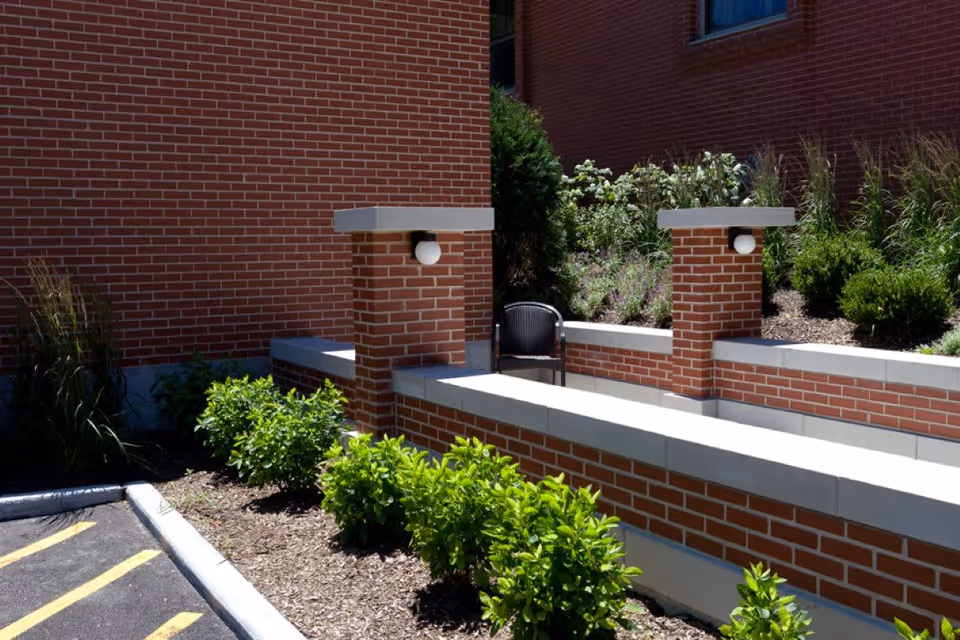 Outdoor seating area with brick walls and pillars, surrounded by green shrubs and plants, next to a parking lot with yellow lines.