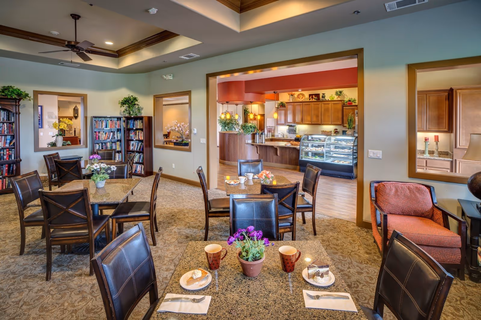 A cozy dining area in Eskaton Village Roseville featuring several tables with dark leather chairs. On the tables are plates with desserts, cups, and small flower pots. The room has carpeted floors, bookshelves filled with books, and large windows looking into an adjacent kitchen area with wooden cabinets and a display case filled with pastries. The ceiling has recessed lighting and a ceiling fan, and there is a comfortable armchair with red upholstery near a side table and lamp.