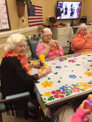Three elderly women sitting around a table decorated with a colorful floral tablecloth. They are wearing bright flower leis and appear to be enjoying a social gathering. An American flag and a TV screen showing people dancing are visible in the background.