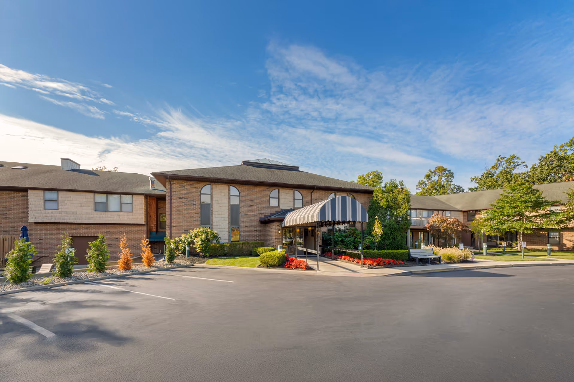 Exterior view of a senior living facility building with a striped awning over the entrance, surrounded by landscaped greenery and a parking lot in front under a partly cloudy blue sky.