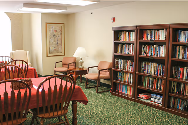 A cozy room with a green patterned carpet, wooden bookshelves filled with books, a table covered with a red tablecloth surrounded by wooden chairs, two armchairs with a small table and lamp between them, and a framed artwork on the wall.
