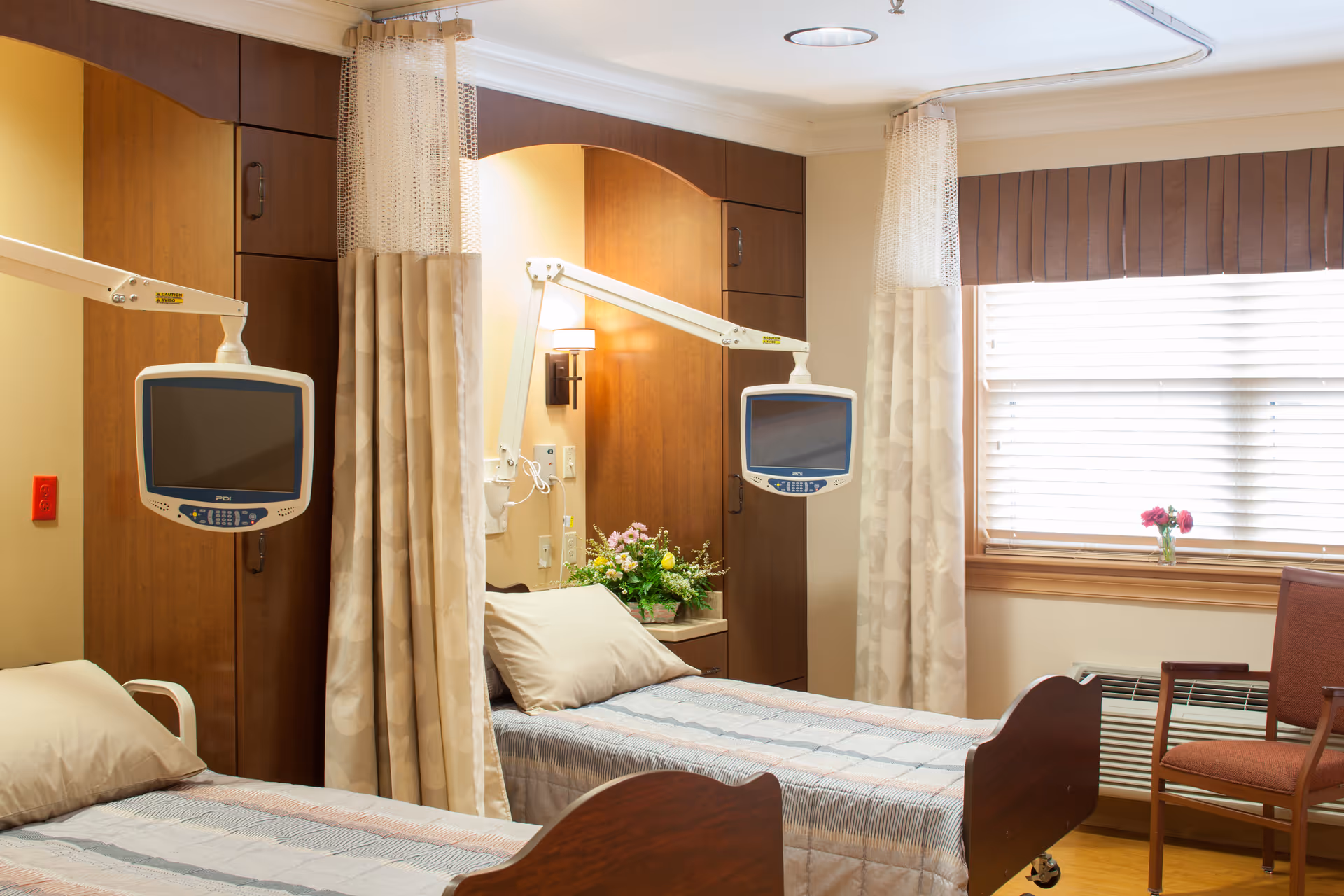 A well-lit hospital or care facility room with two beds separated by a privacy curtain. Each bed has a mounted television screen on an adjustable arm. There is a window with blinds and a small vase with flowers on the windowsill. A wooden chair is placed near the window, and a floral arrangement is on a small table between the beds.