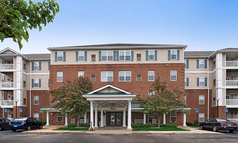 Front exterior view of The Meadows of Brownstown senior living facility, featuring a multi-story brick and siding building with a covered entrance supported by white columns, surrounded by trees and parked cars.