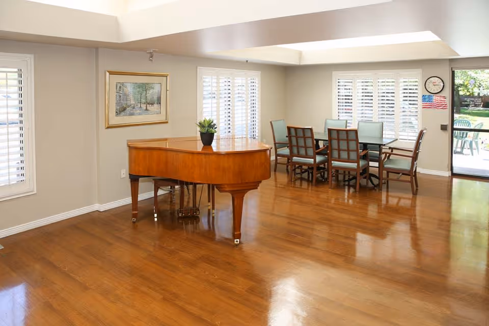 A bright room with wooden flooring featuring a grand piano with a small plant on top, a dining table with six chairs, large windows with white shutters, a framed painting on the wall, a clock, and an American flag near a glass door leading outside to a patio area.