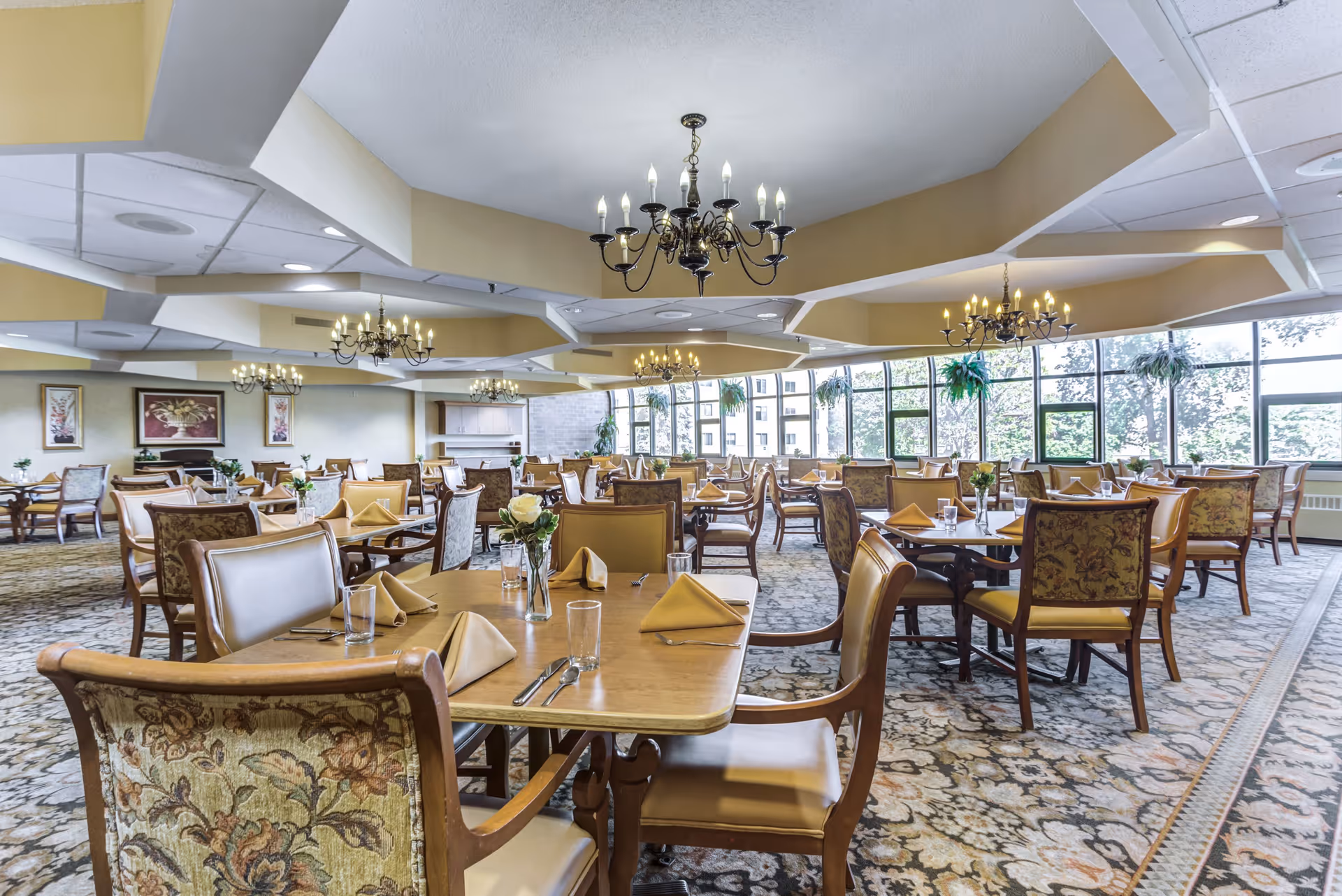 A spacious dining room with multiple wooden tables and chairs arranged neatly. Each table is set with folded napkins, glasses, and silverware. The room features large windows allowing natural light to fill the space, chandeliers hanging from the ceiling, and decorative plants near the windows. The carpet has a floral pattern, and framed artwork is visible on the walls.