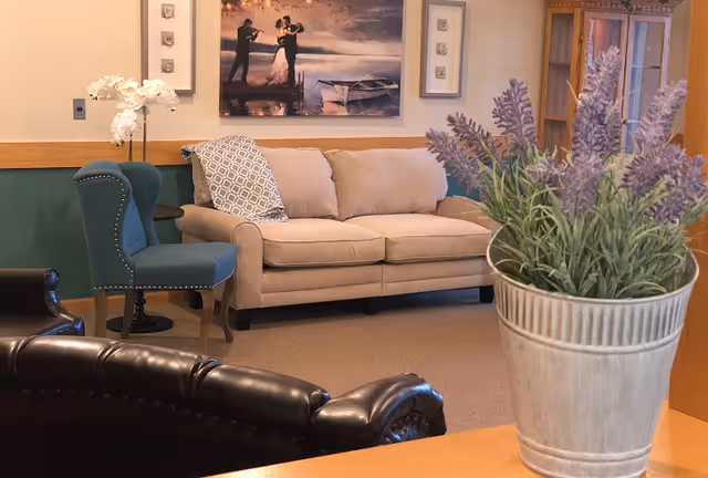 A cozy living room area with a beige sofa, a green upholstered chair with a patterned pillow, a dark brown leather chair, and a potted lavender plant in the foreground. The walls are decorated with framed artwork and a large painting of a couple dancing by the water.