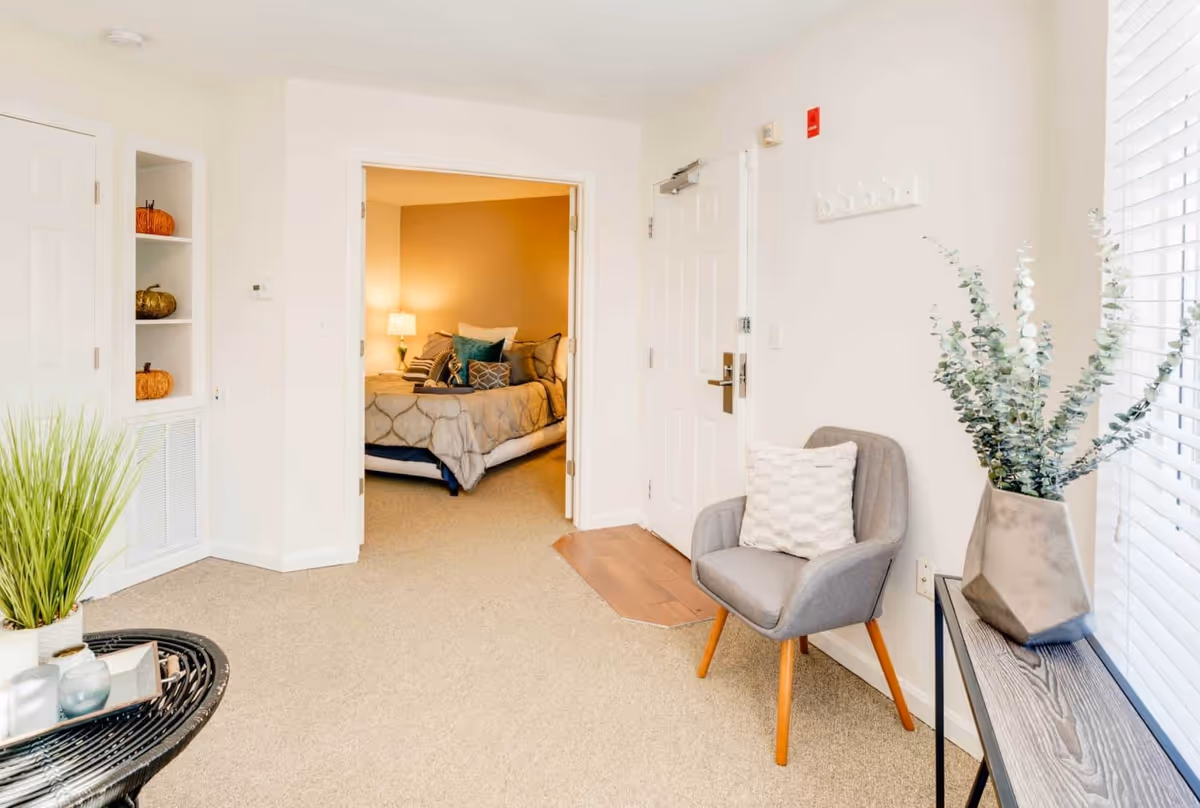 Bright sitting area with a gray armchair, decorative plants, and an open doorway revealing a bed in the adjacent bedroom.