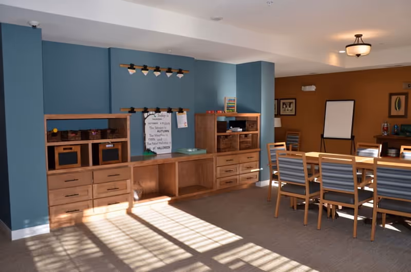 A well-lit room with blue and brown walls featuring wooden cabinets with drawers and shelves. There is a long wooden table with several chairs around it, a whiteboard on a stand, and framed pictures on the walls. Sunlight streams through a window, casting shadows on the carpeted floor.
