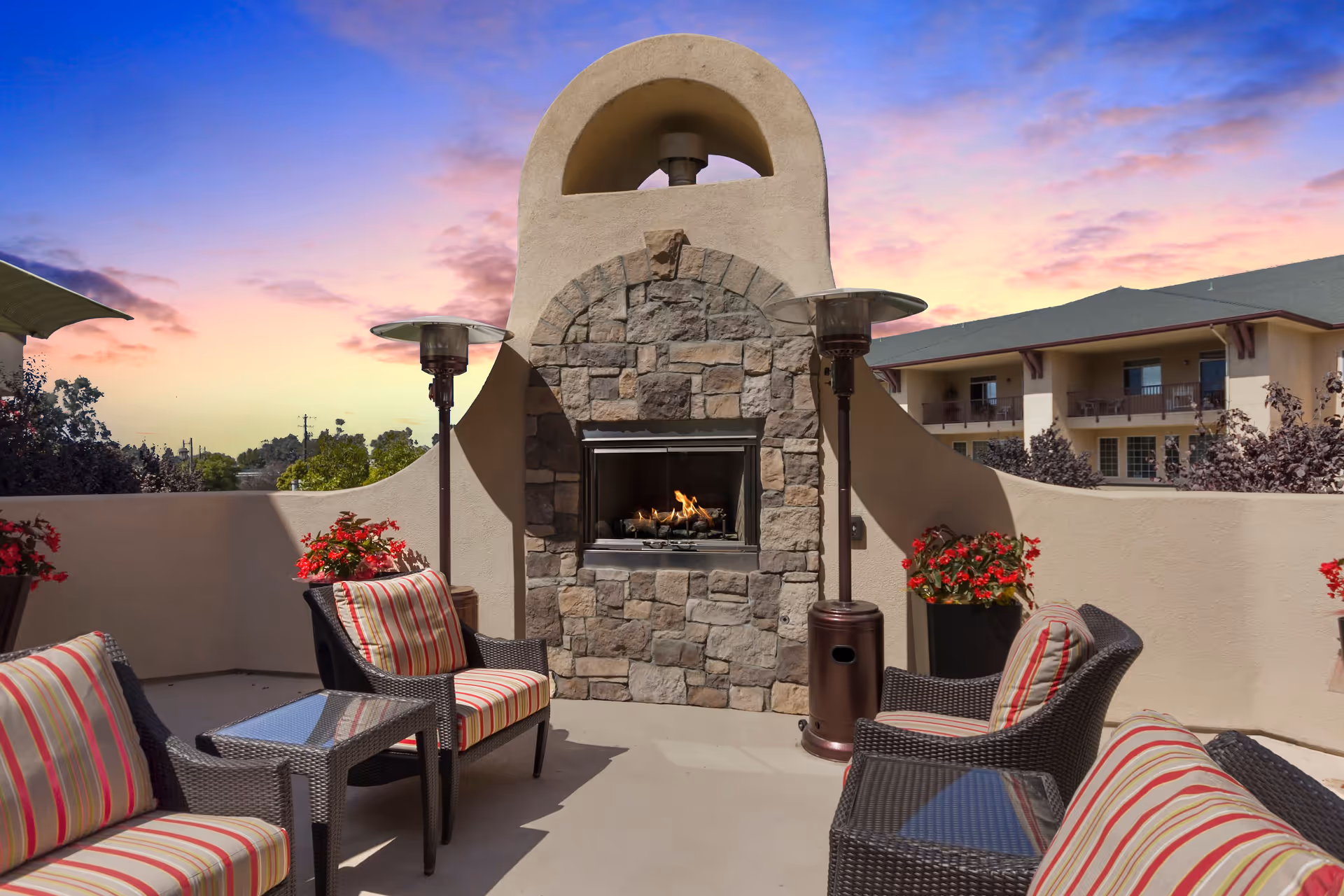 Outdoor patio area with a stone fireplace in the center, surrounded by wicker chairs with striped cushions and glass-top side tables. There are two tall outdoor heaters on either side of the fireplace and potted red flowers along the beige stucco walls. The sky is colorful with a sunset or sunrise.
