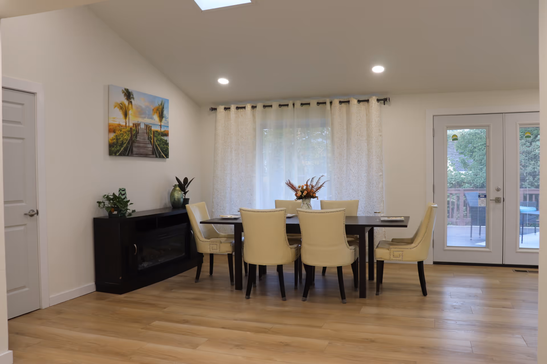 A dining area with a dark wooden table surrounded by six cream-colored upholstered chairs. Behind the table is a window with sheer white curtains, and to the right are glass double doors leading to an outdoor deck. On the left wall, there is a painting of a boardwalk leading to a beach with palm trees, and below it is a black cabinet with plants and decorative items. The floor is light wood, and the ceiling has recessed lighting and a skylight.