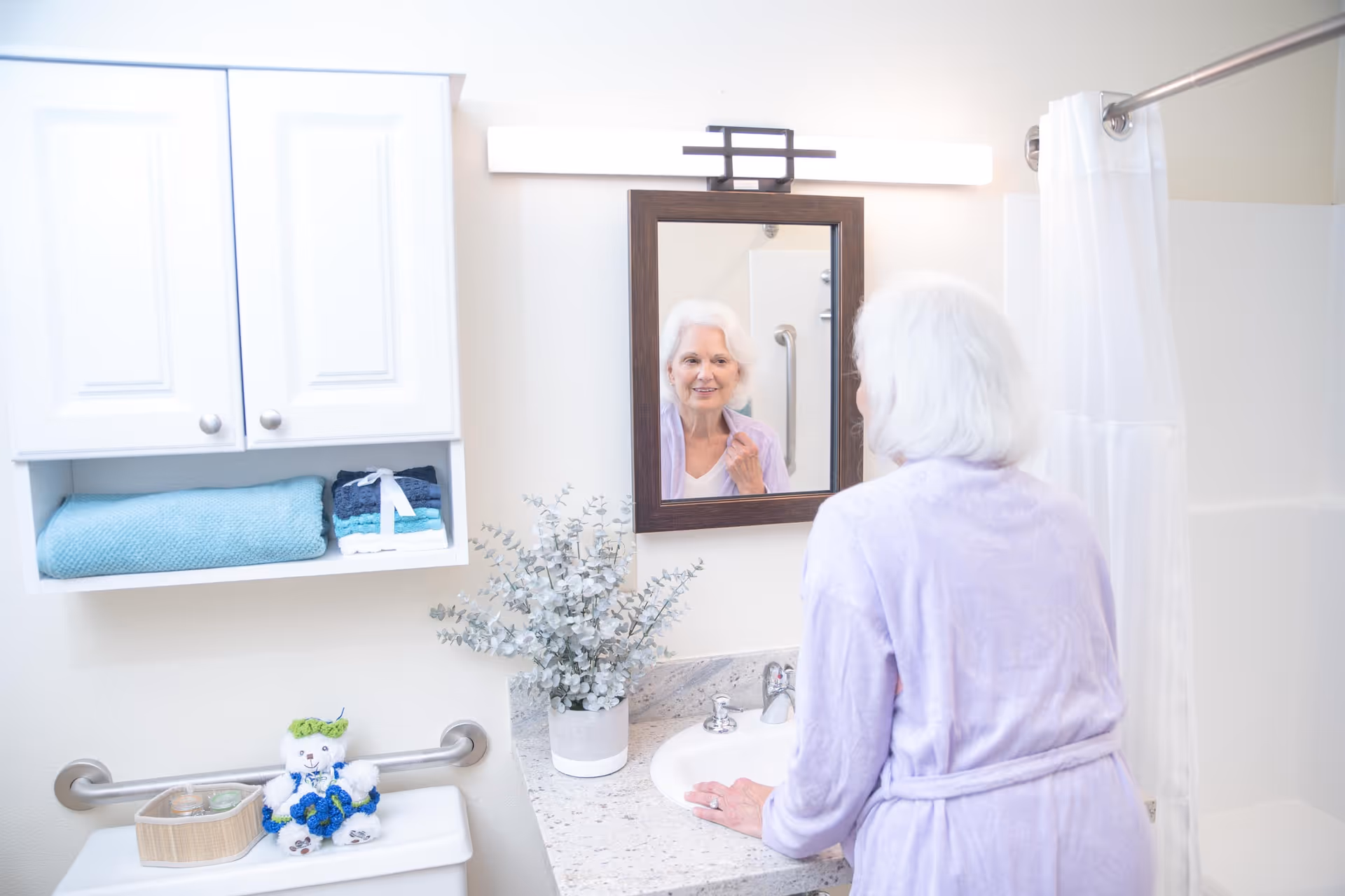 An elderly woman with white hair wearing a lavender robe is standing in a bathroom, looking at herself in a mirror above a sink. The bathroom has a white cabinet with folded towels, a small potted plant on the countertop, a shower with a white curtain, and a grab bar next to the toilet which has a small teddy bear and a basket on top.