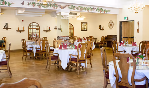 A spacious dining room with multiple round tables covered with white tablecloths and set with folded red napkins and small floral centerpieces. The room features wooden chairs, hardwood floors, wall decorations including mirrors and shelves, and ceiling fans with lights. There is an exit door visible in the background.