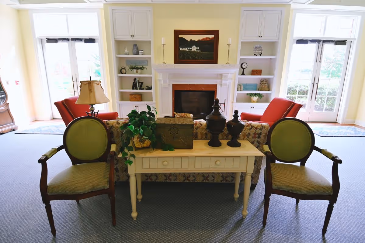 A cozy living room area with two green cushioned wooden chairs in the foreground, a small white table with decorative items and a plant, a patterned sofa, two red armchairs, built-in white shelves on either side of a white fireplace with a framed painting above it, and large glass doors letting in natural light.