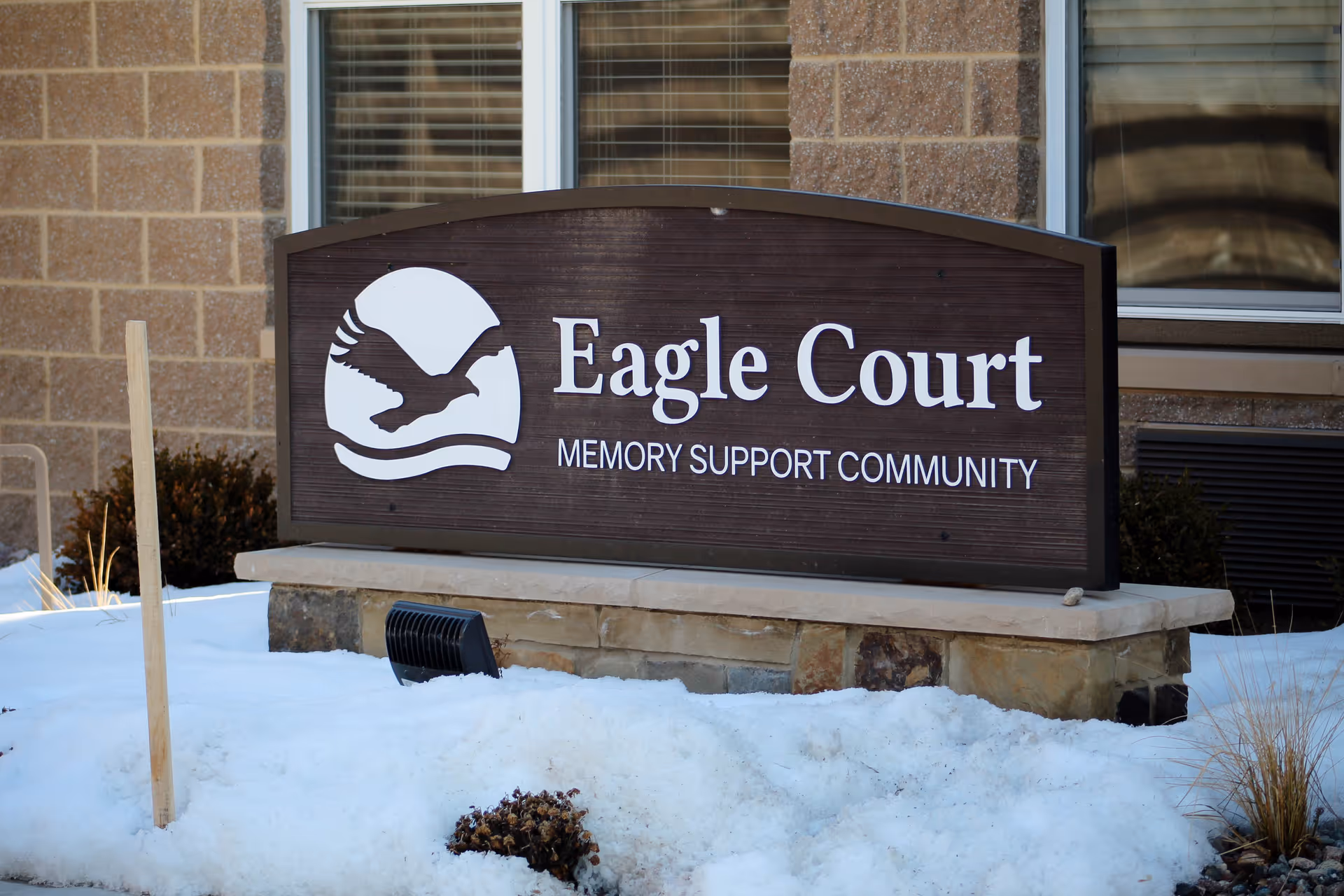 Outdoor sign for Eagle Court Memory Support Community mounted on a stone base with snow on the ground around it, in front of a brick building with windows.